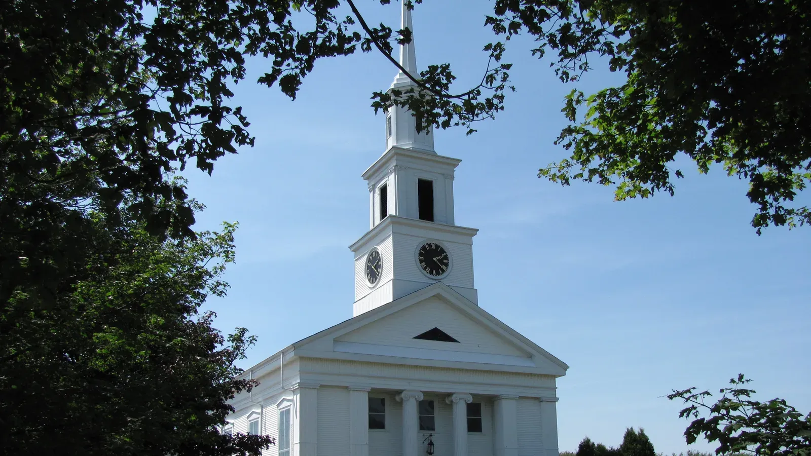 Avon Baptist Church, a white Greek Revival meetinghouse with a clock tower and spire framed by summer trees, Avon, Massachusetts