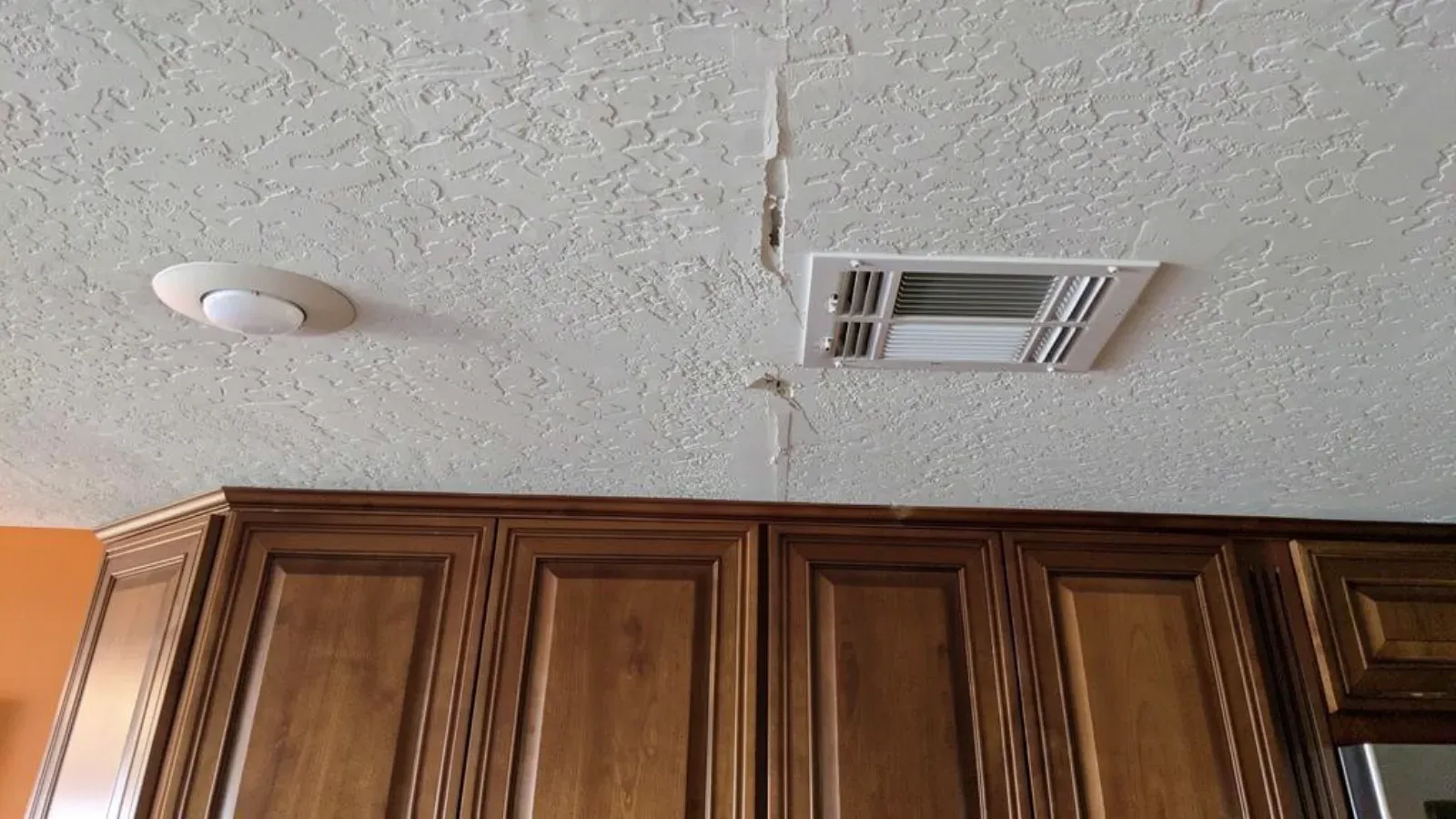 A crack running through a textured kitchen ceiling beside a white rectangular ceiling air register, the paint and joint tape split open along its length, above stained wood upper cabinets