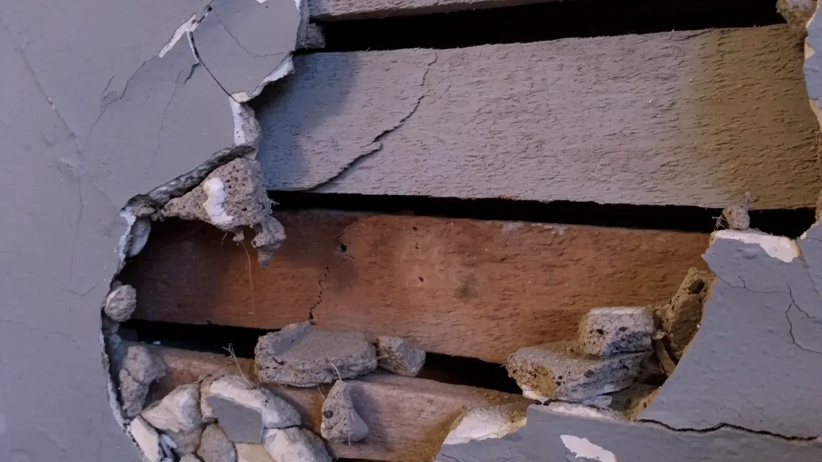Hole broken through a painted plaster wall, exposing the horizontal wood lath behind it and the crumbling plaster keys around the opening